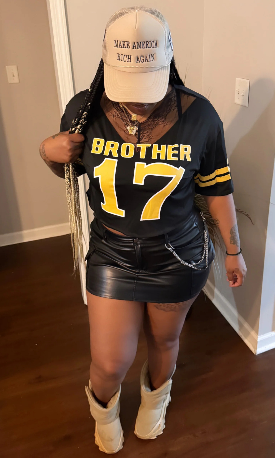 Model wearing black “Brother 17” graphic jersey top with yellow print, paired with black leather mini skirt, beige boots, and beige cap indoors.