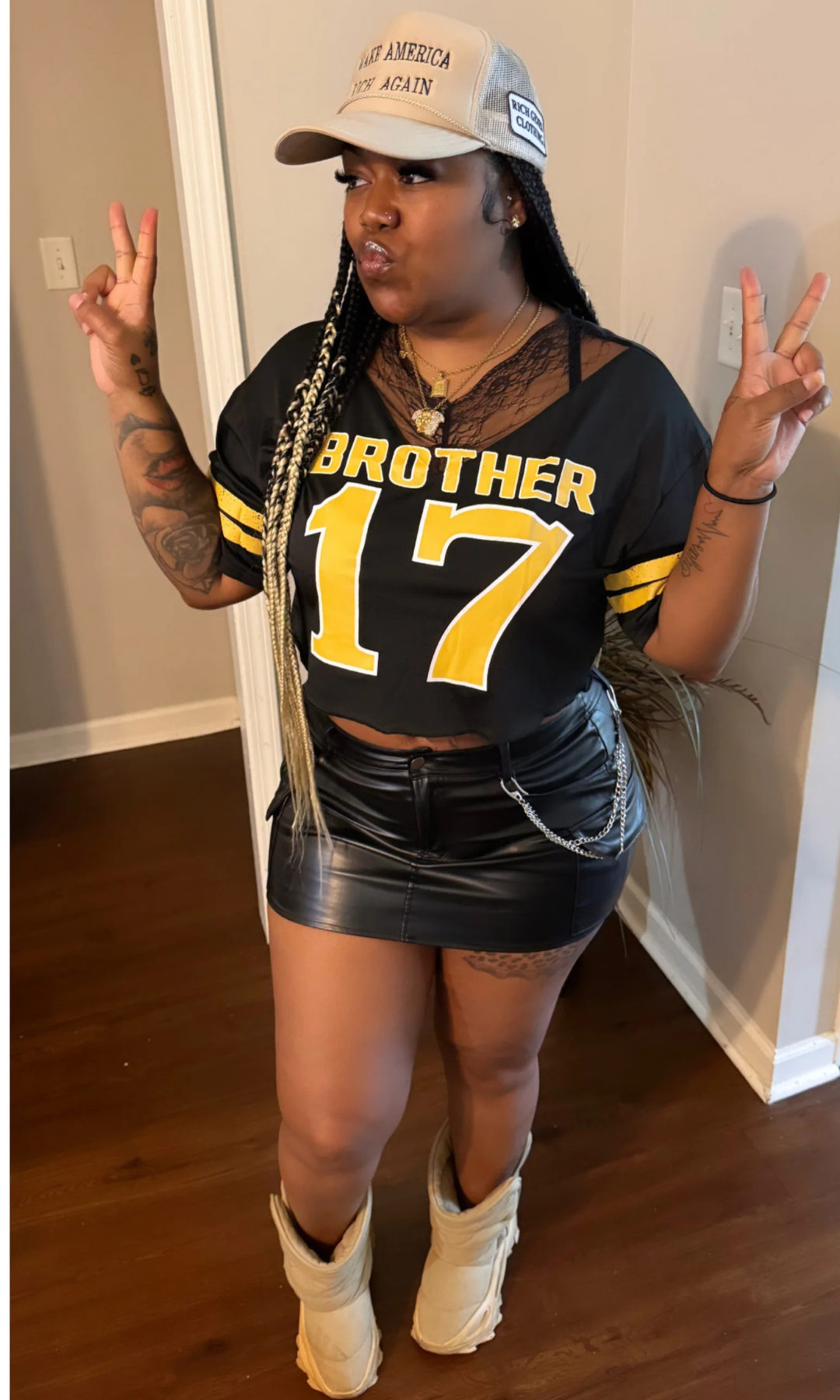 Front pose of model in black “Brother 17” jersey top with yellow lettering, black leather skirt, and beige cap, making peace signs.