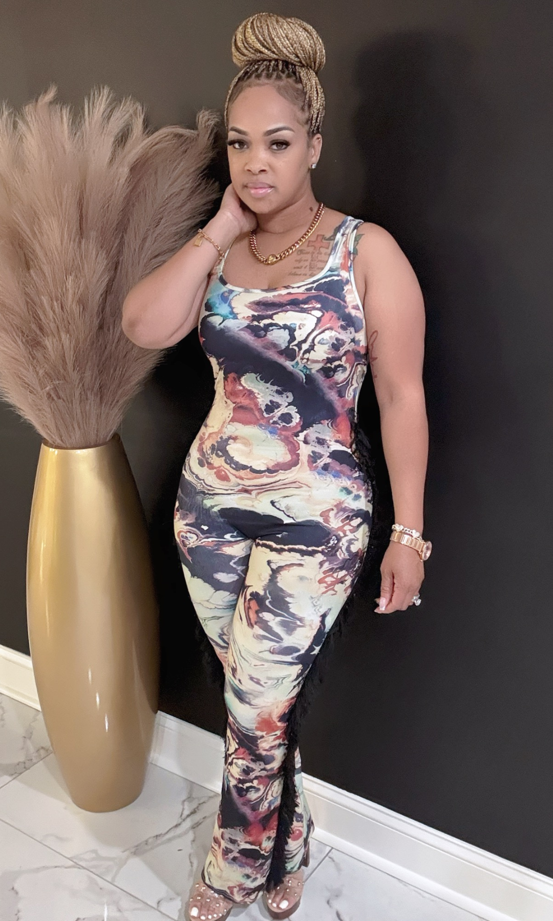 Woman in a colorful jumpsuit standing next to a decorative vase against a black wall.