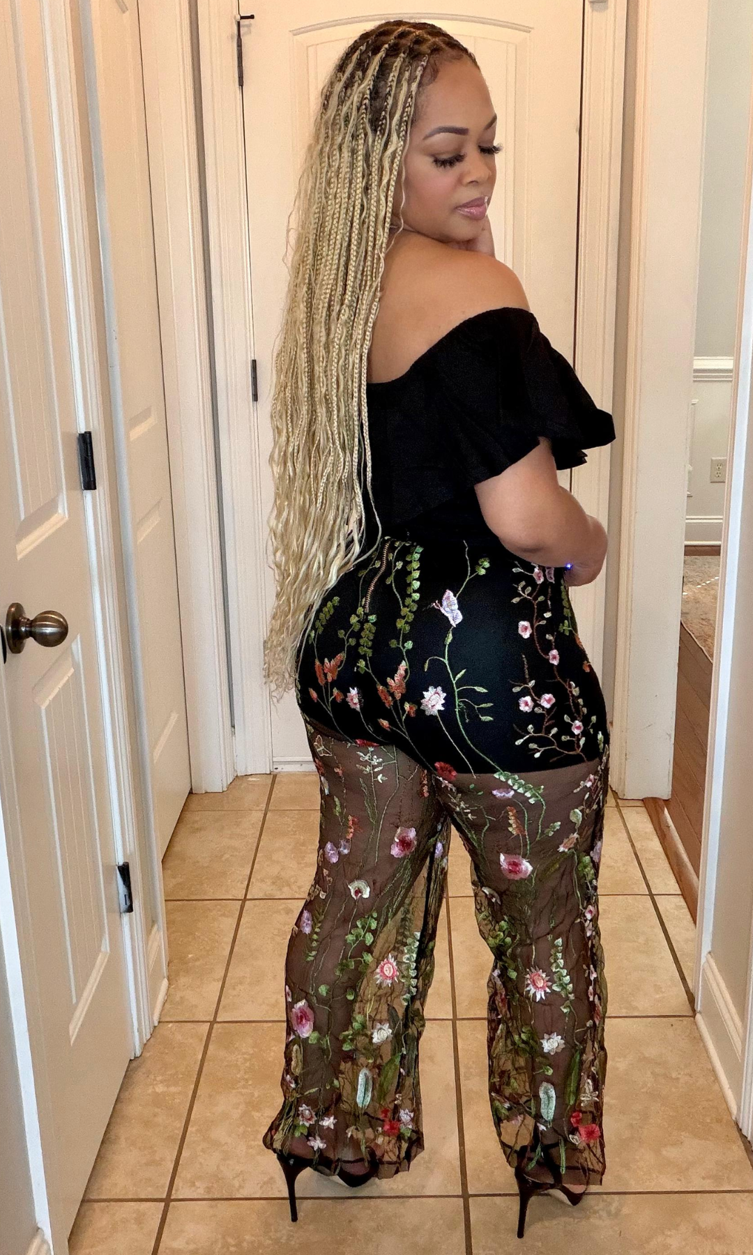 Woman wearing a black off-shoulder top and floral sheer pants in a room.