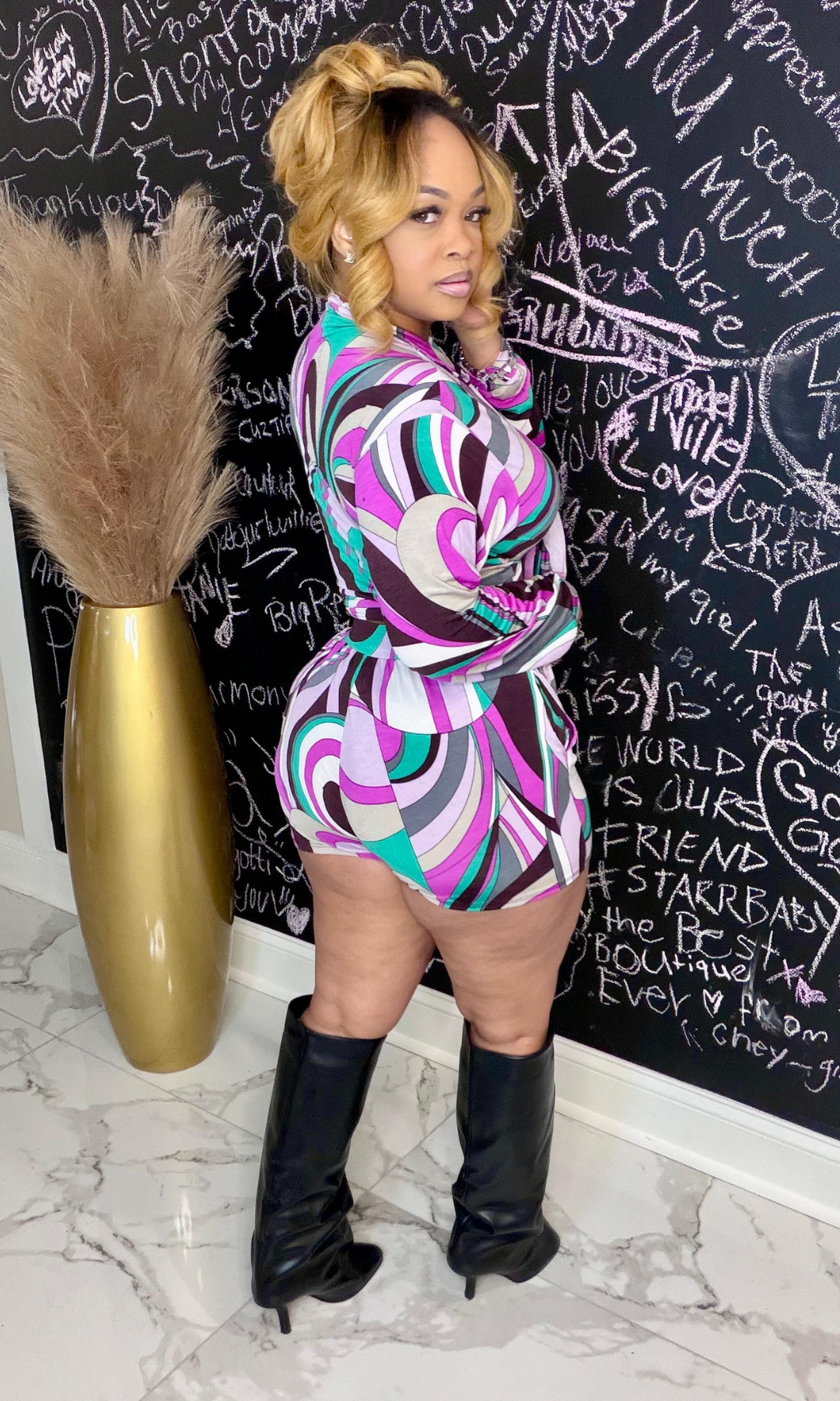 Person wearing a colorful patterned outfit with black knee-high boots in front of a chalkboard wall.