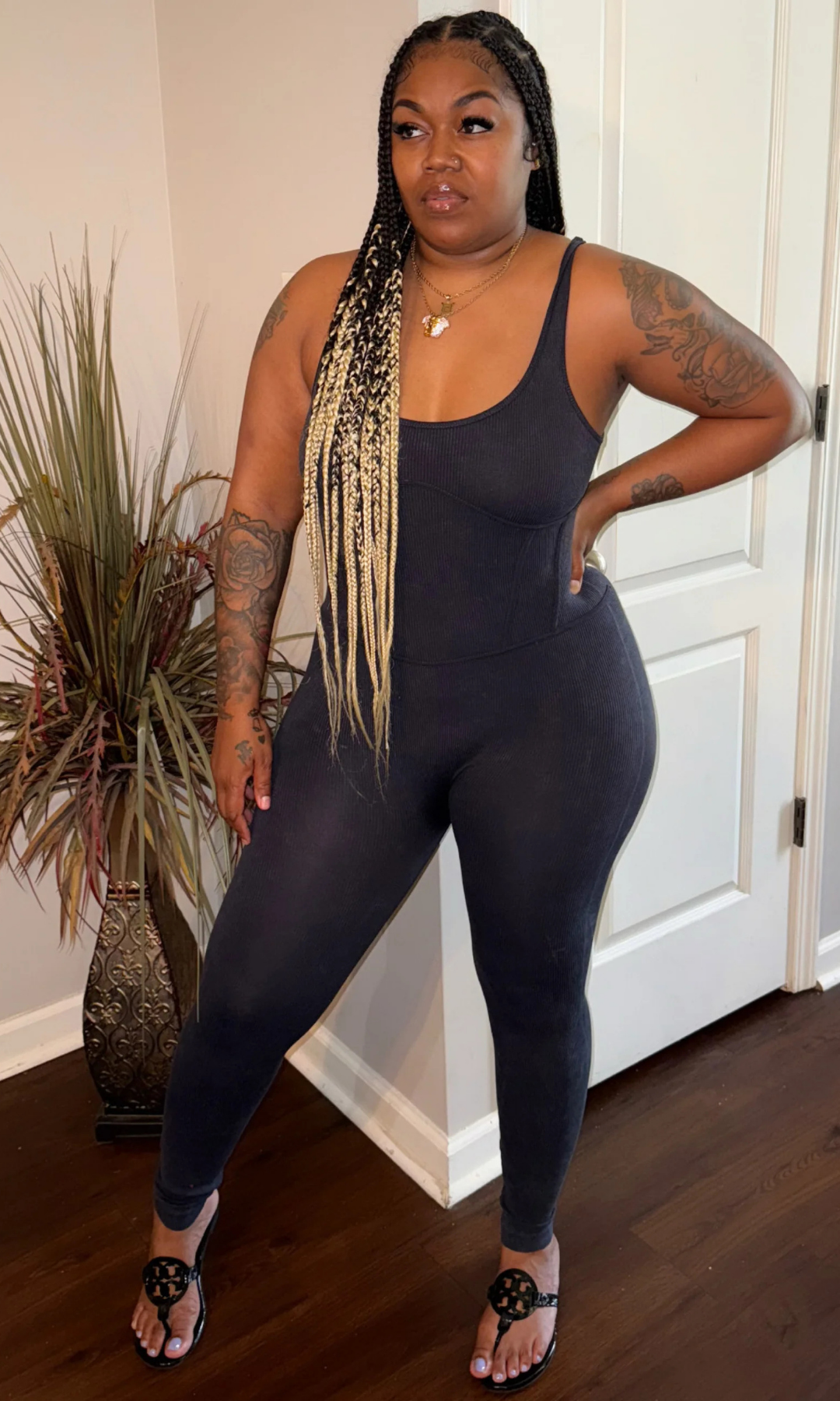 Front view of woman wearing a black, sleeveless, fitted Tiff Tiff jumpsuit with scoop neckline, styled with black sandals and gold layered necklaces, standing in a modern indoor setting.