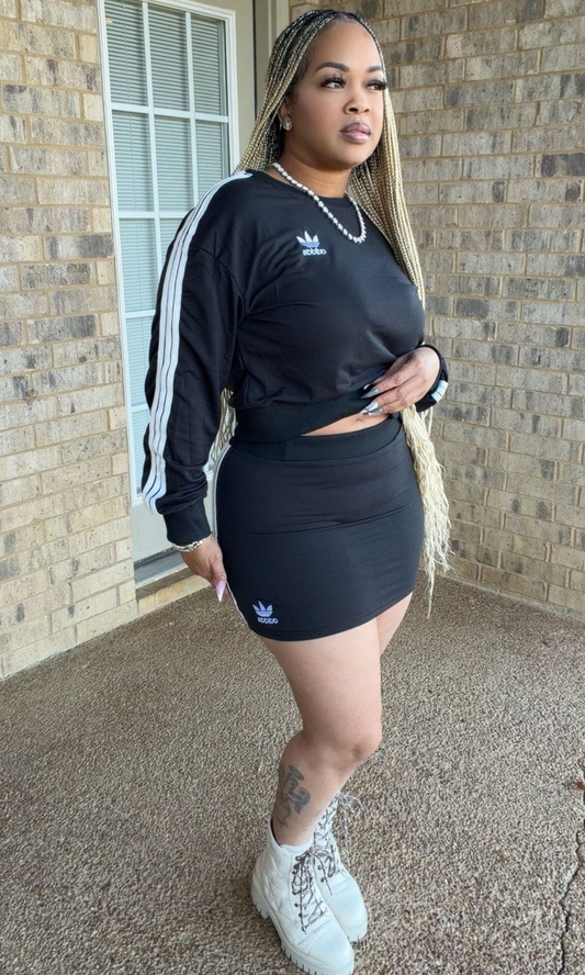 A woman modeling the Adi-Das Skirt Set in black, featuring a long-sleeve athletic-inspired pullover with white sleeve stripes and a matching high-waisted fitted mini skirt, styled with lace-up boots.