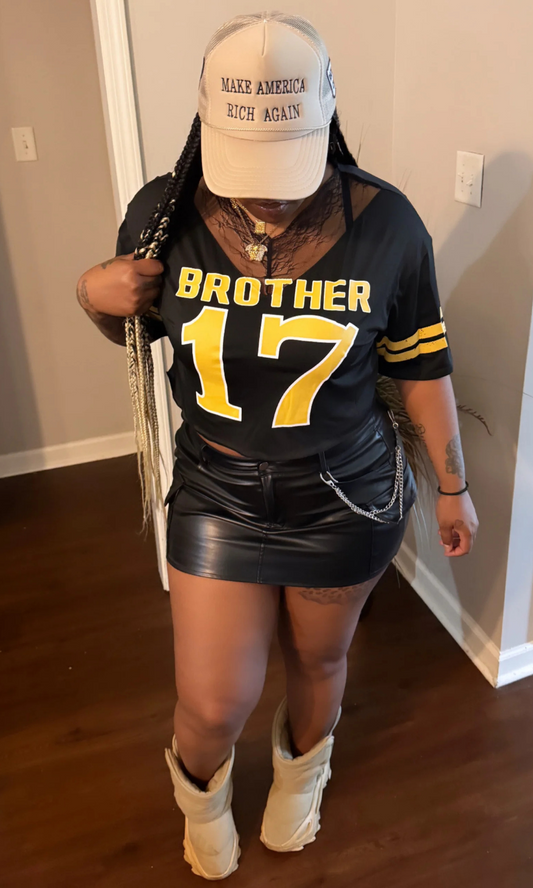 Model wearing black “Brother 17” graphic jersey top with yellow print, paired with black leather mini skirt, beige boots, and beige cap indoors.