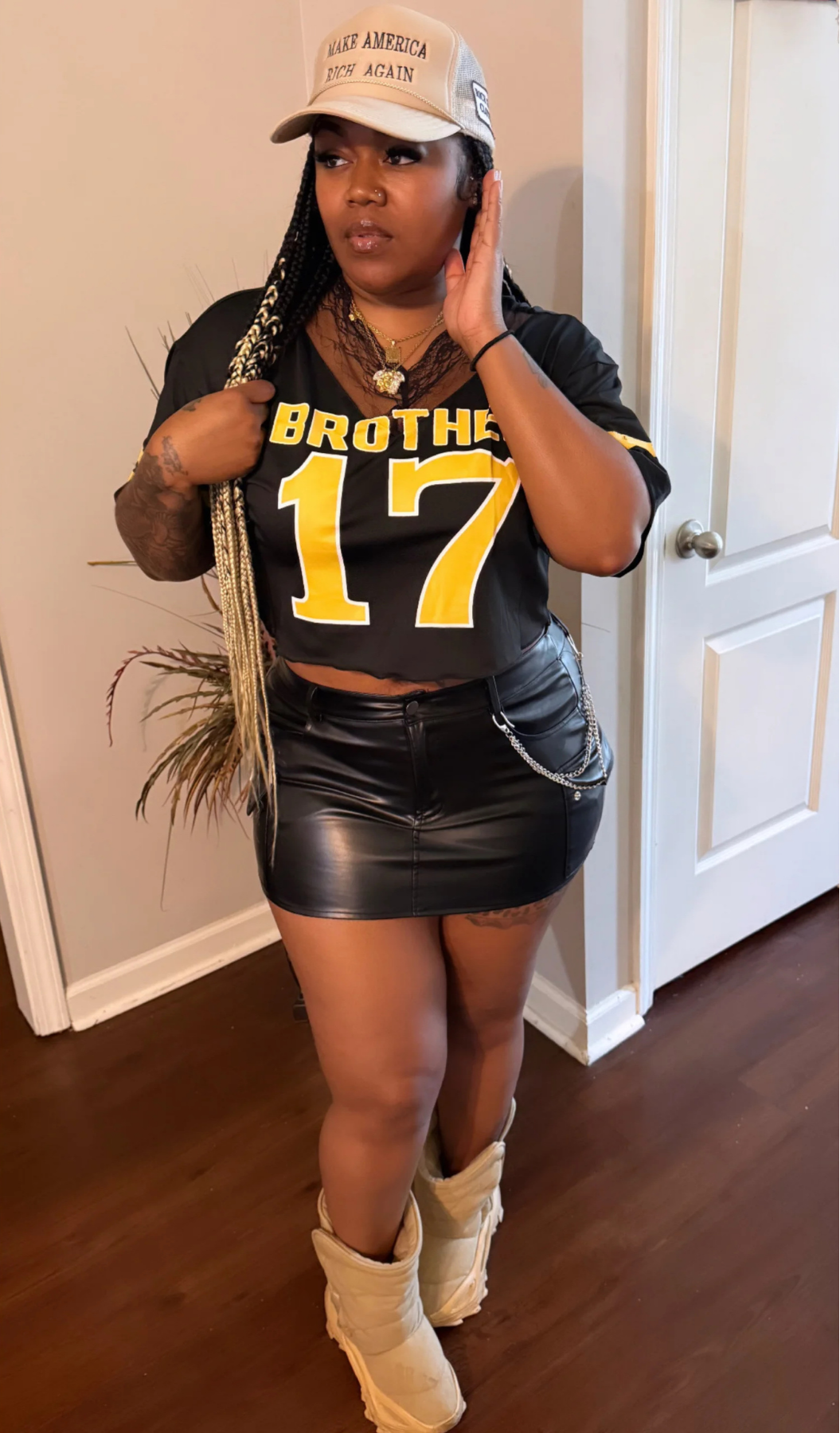 Model in black “Brother 17” jersey top styled with black leather mini skirt, chain detail, beige boots, and beige cap, standing indoors.