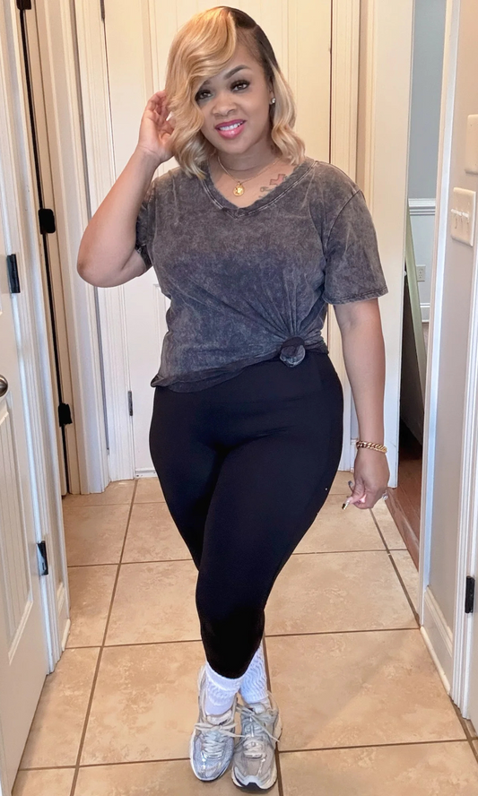 Model in washed black oversized top knotted at the waist, black leggings, white socks, and silver sneakers indoors.