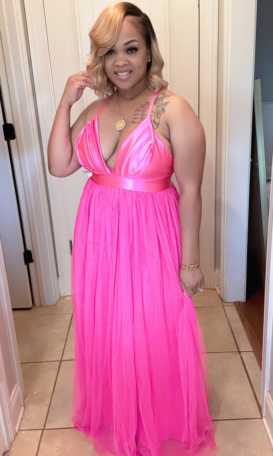 Woman wearing a vibrant Barbie Pink Maxi Dress with plunging neckline and satin waistband, standing in a doorway.