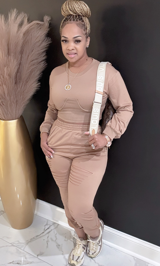 Woman in a beige outfit standing next to a decorative vase with pampas grass against a black wall.
