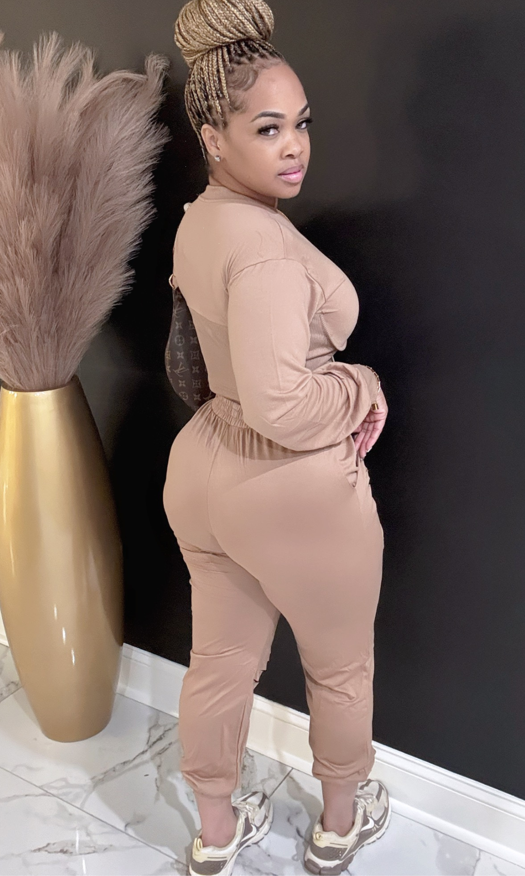 Woman in a beige jumpsuit standing against a dark wall with decorative elements.