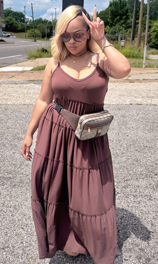 Woman in a long brown dress standing on a street, making a peace sign.