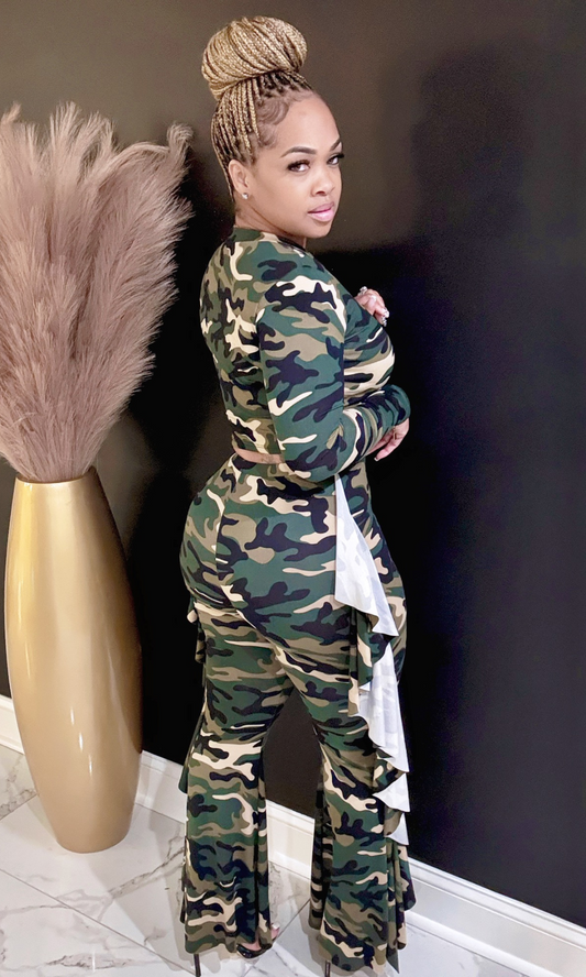 Woman in a camouflage outfit standing next to a decorative vase against a dark wall.