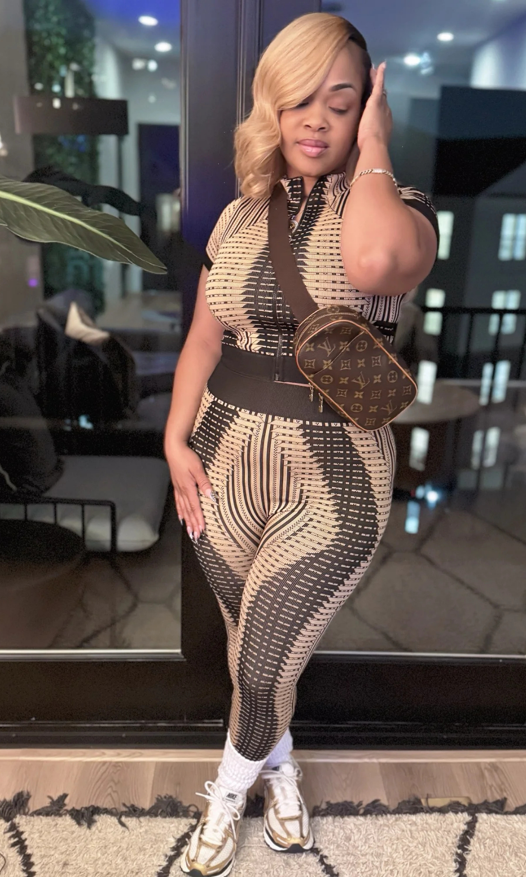 Woman in a patterned outfit with a handbag standing in a modern indoor setting.