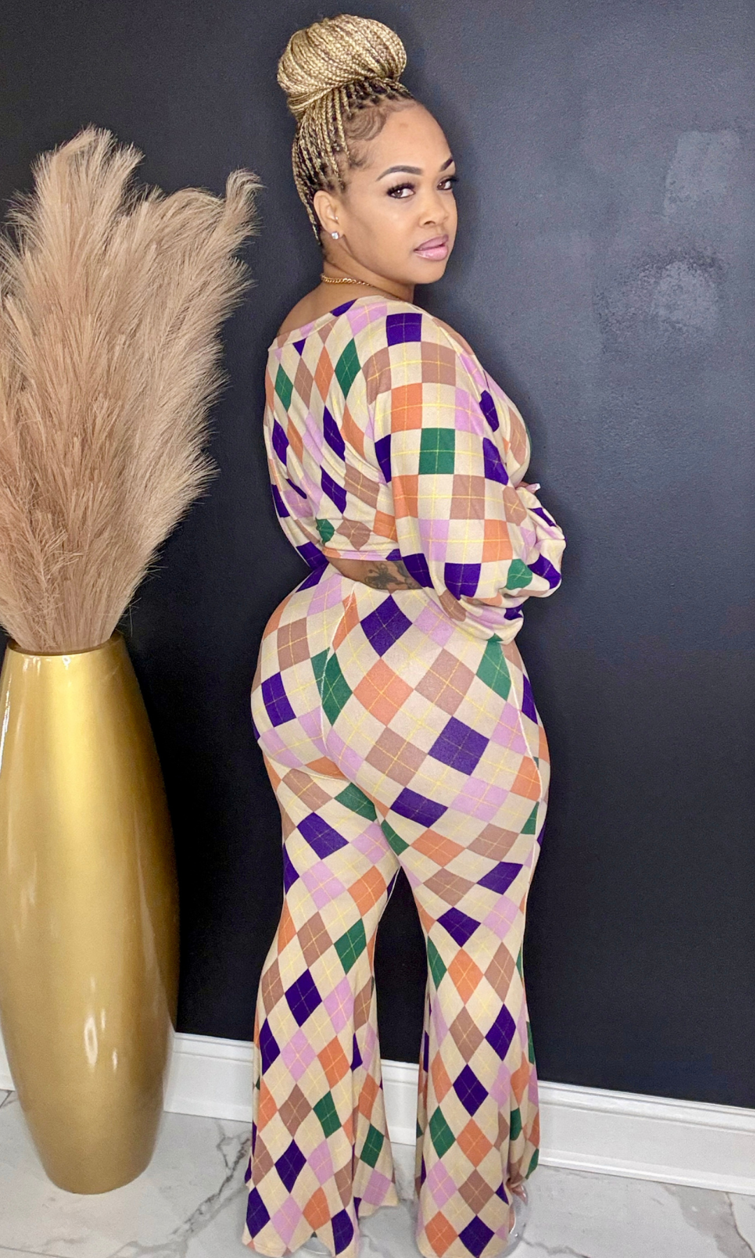 Woman wearing a colorful checkered jumpsuit standing against a dark wall with a gold vase and dried plants.