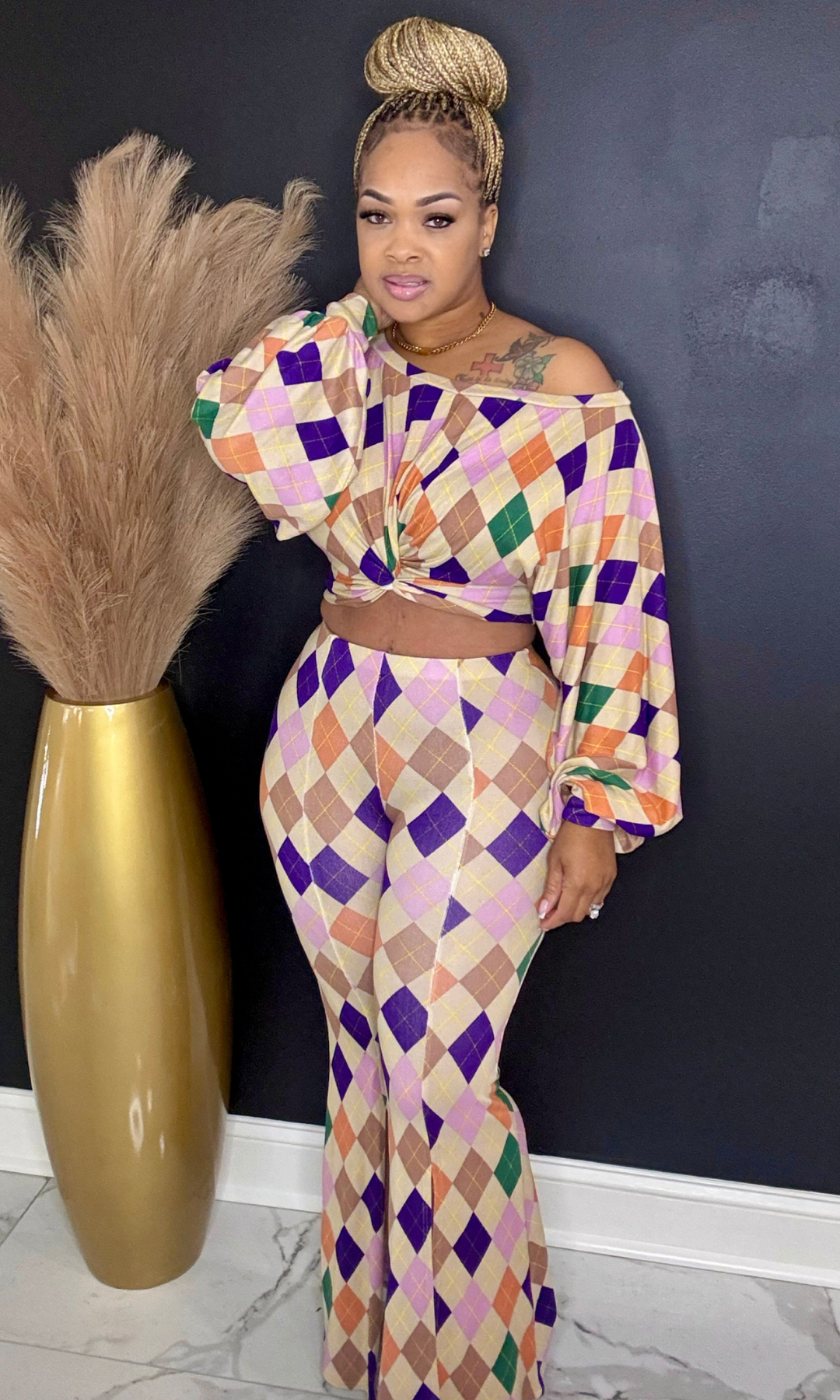 Woman wearing a colorful geometric outfit standing next to a gold vase against a dark wall.
