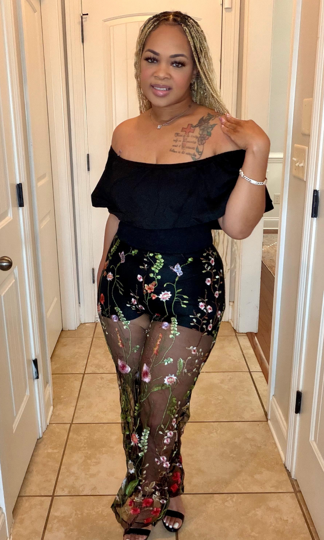 Woman wearing a black off-shoulder top and floral sheer pants in a hallway.
