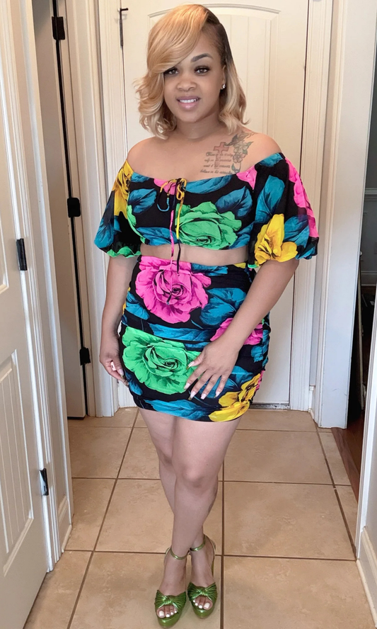 Woman wearing a colorful floral dress standing in a room with white walls and a tiled floor.