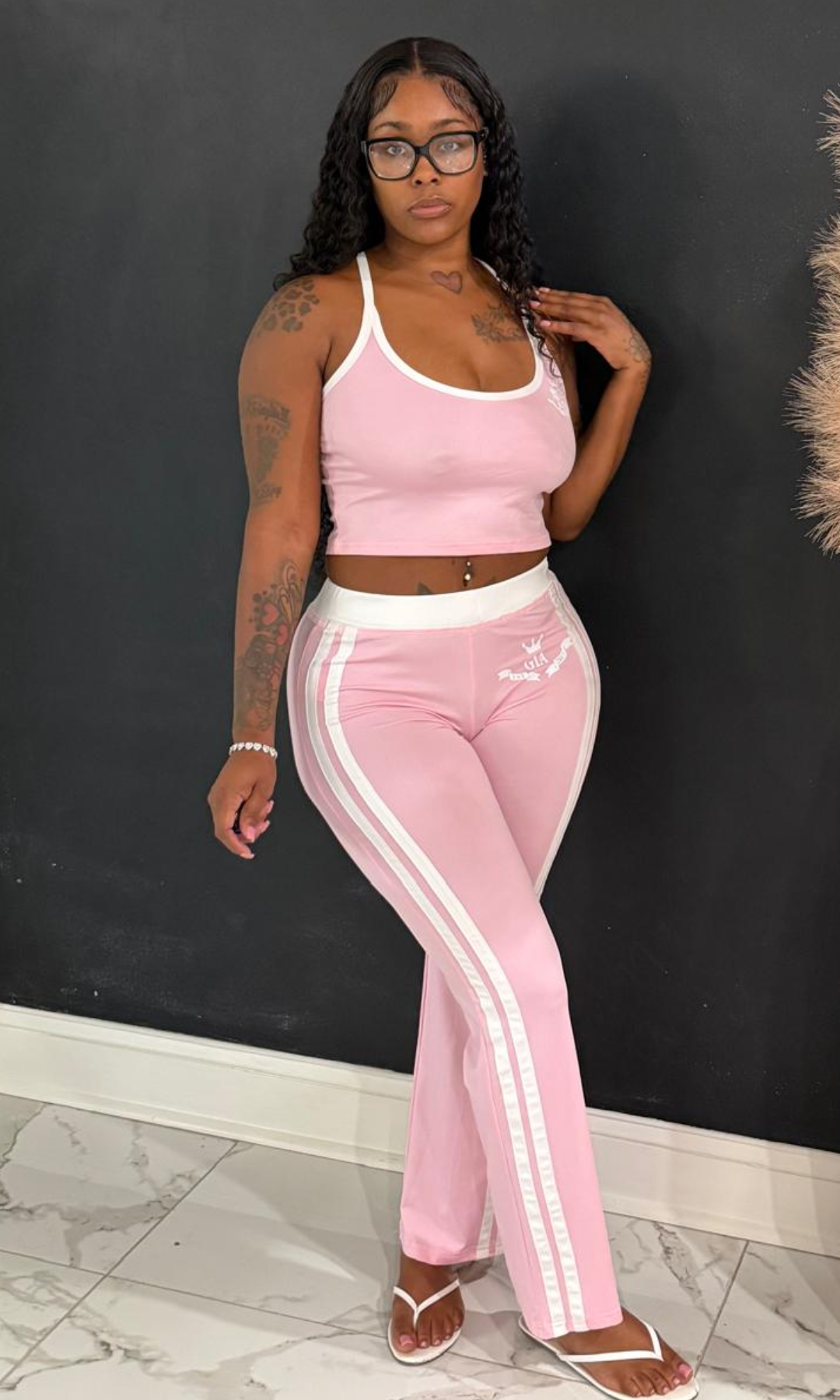 A woman modeling a pink two-piece tracksuit with white trim, consisting of a cropped tank top and matching track pants with white side stripes, standing against a dark gray wall.