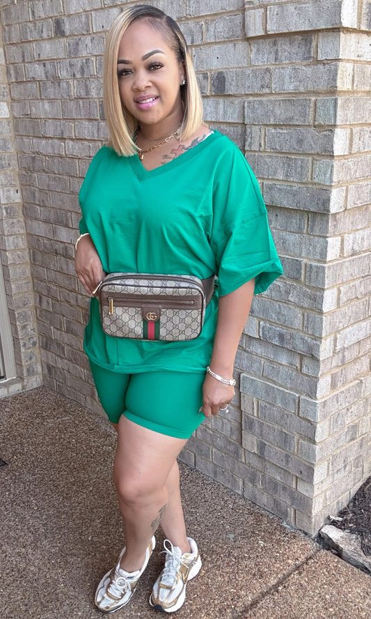 Woman in green outfit with a patterned bag standing against a brick wall.