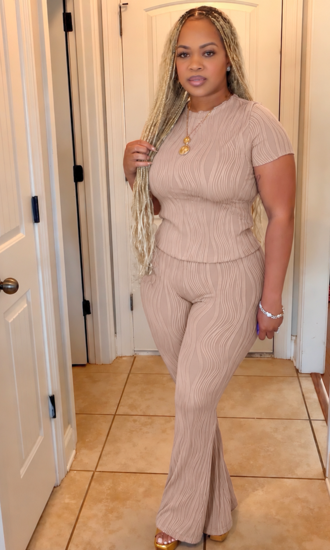 A woman modeling a ribbed two-piece jumpsuit in a light beige or taupe color, standing in a tiled hallway with white doors.