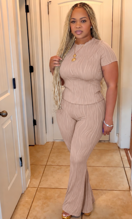 A woman modeling a ribbed two-piece jumpsuit in a light beige or taupe color, standing in a tiled hallway with white doors.