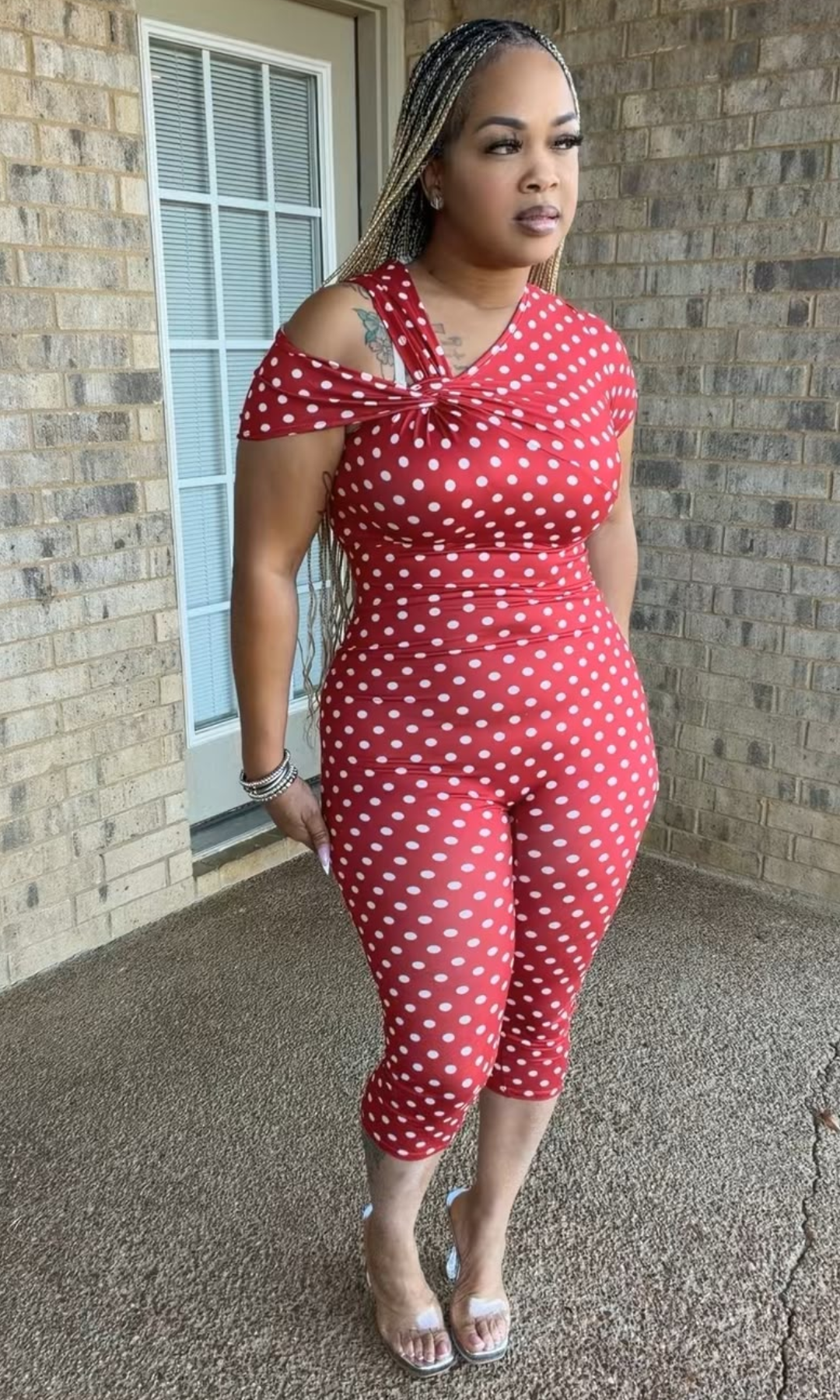Woman modeling the Red Polka Babe Jumpsuit featuring a vibrant red and white polka dot print, asymmetrical twisted neckline, off-shoulder detail, and fitted capri-length bottoms.