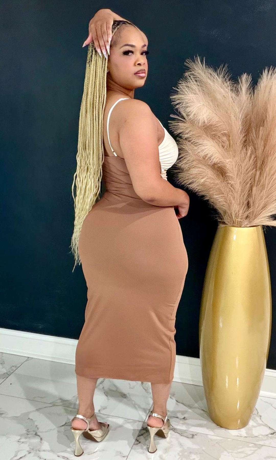 A woman modeling a fitted tan midi dress with a white bandeau-style top, standing indoors beside a tall gold vase with pampas grass against a dark blue wall.
