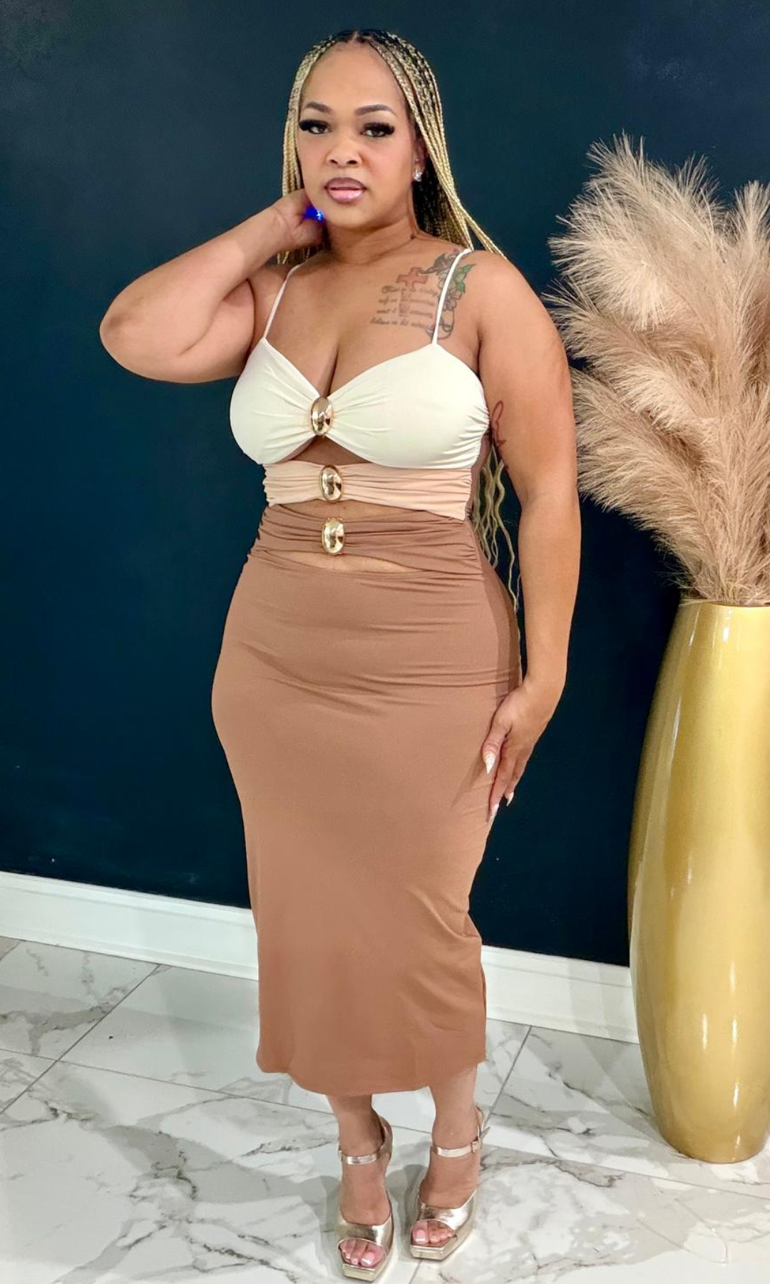 A woman models a two-piece dress set featuring a cream-colored sweetheart bustier top with gold buttons and a matching beige ruched midi skirt with gold button accents, paired with clear high-heeled sandals.