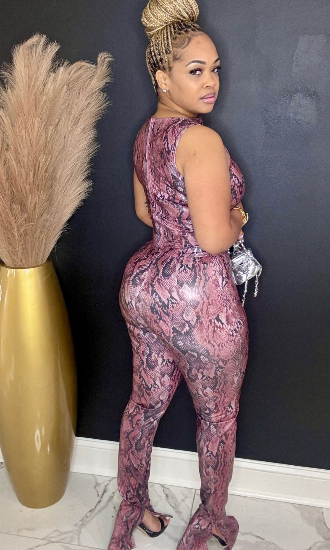 Woman wearing a pink snake print jumpsuit standing against a dark wall with decorative elements.