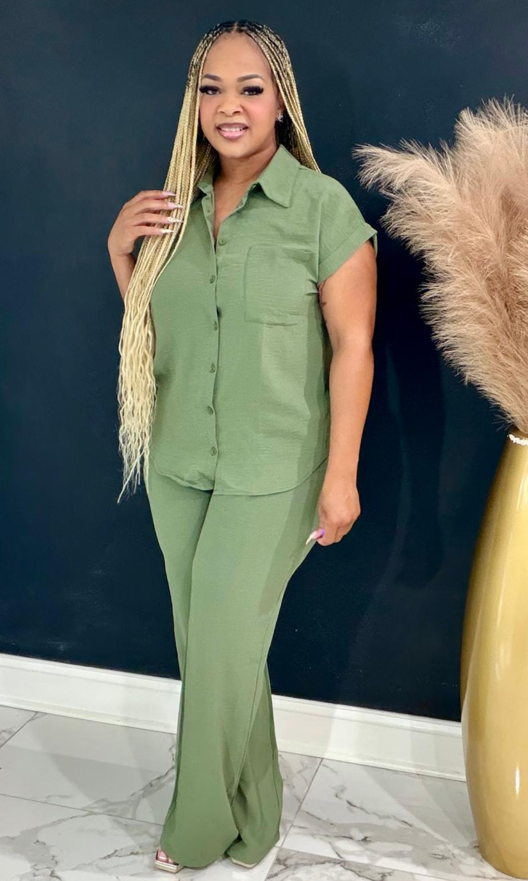 A woman modeling a matching sage green two-piece set consisting of a short-sleeve button-up shirt and wide-leg pants, standing in front of a dark blue wall with a decorative gold vase and pampas grass.