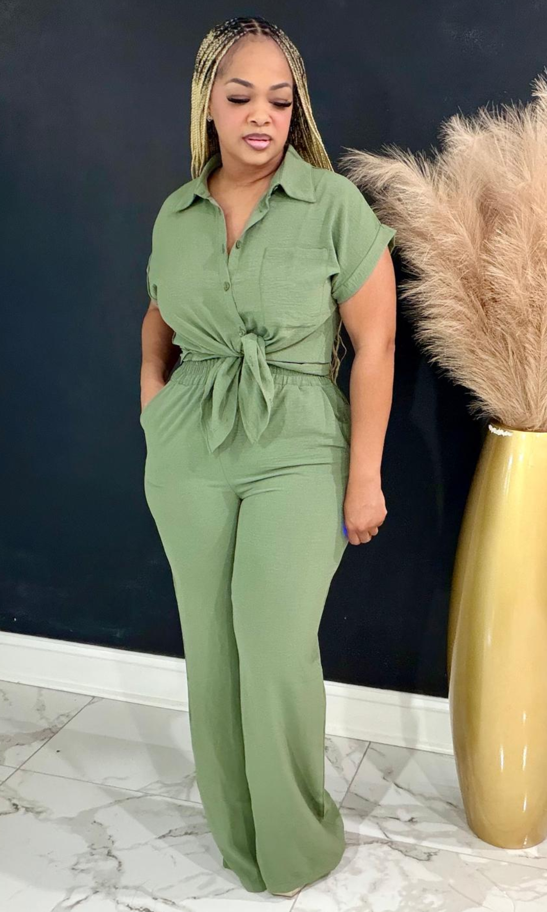 A woman modeling a sage green short-sleeve jumpsuit with a tie-front waist, standing indoors against a dark wall next to a gold vase with dried pampas grass.