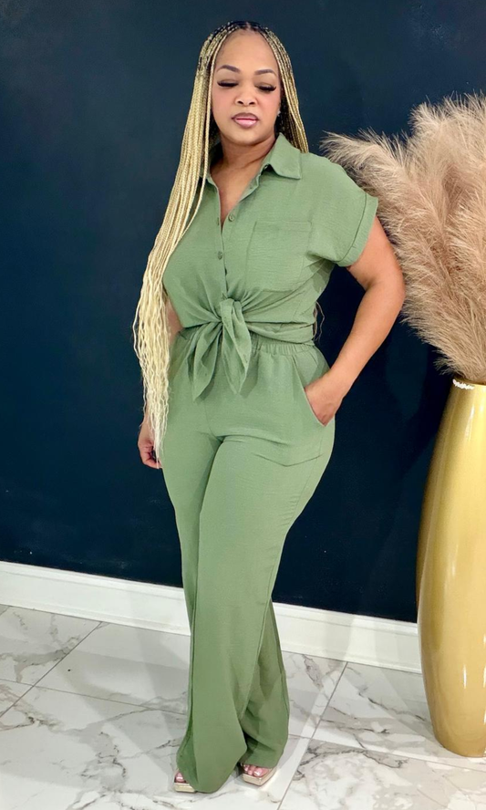 A woman modeling a sage green short-sleeve jumpsuit with a front knot detail at the waist and side pockets, standing in an indoor setting with a dark blue wall and a gold vase with pampas grass.