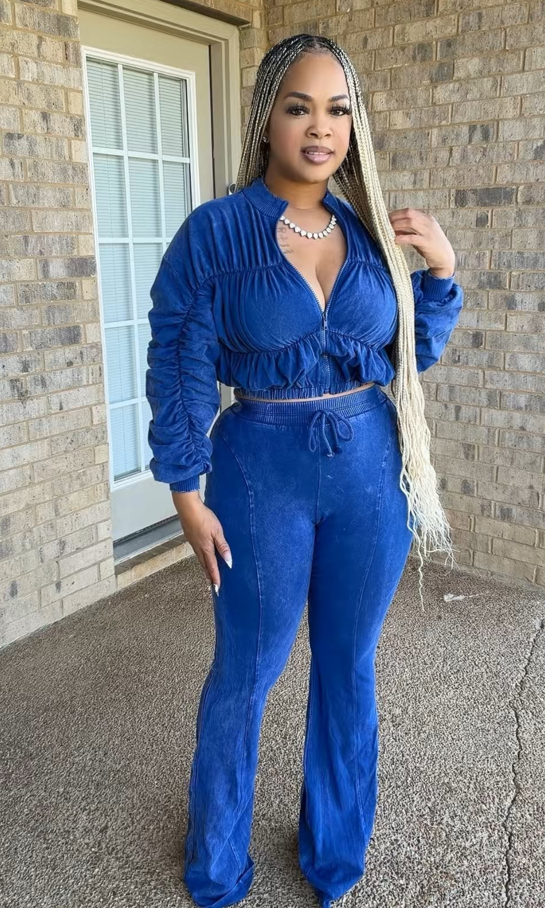 A woman modeling a matching royal blue two-piece lounge set consisting of a long-sleeve ruched crop top and high-waisted flared pants, standing on a concrete porch in front of a brick wall.