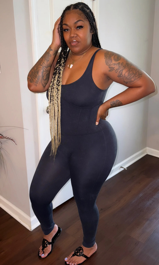 Angled Front view of woman wearing a black, sleeveless, fitted Tiff Tiff jumpsuit with scoop neckline, styled with black sandals and gold layered necklaces, standing in a modern indoor setting.

