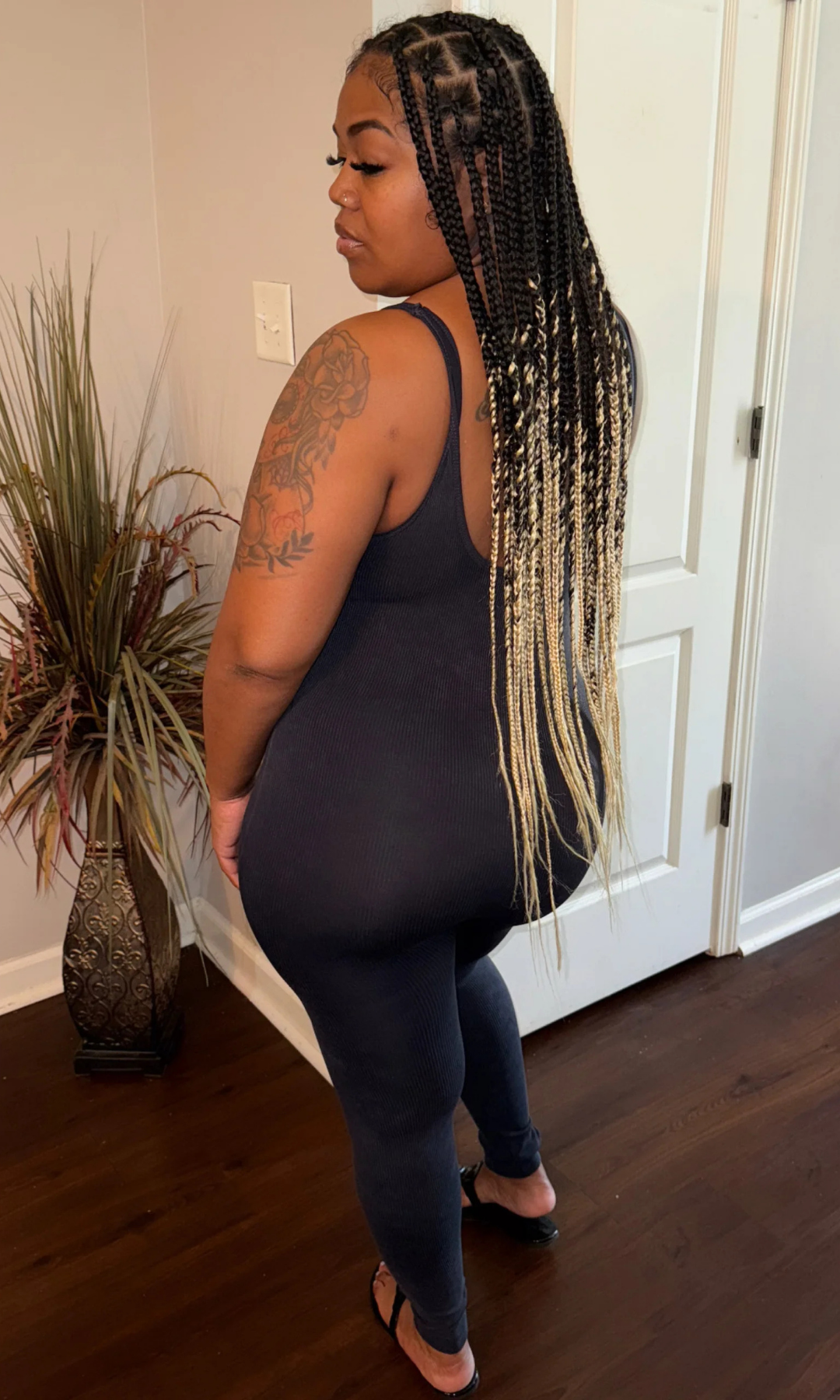 Side and partial back view of woman in a black, sleeveless, fitted Tiff Tiff jumpsuit, showcasing long blonde and black braids, detailed tattoos, and figure-hugging stretch fabric.

