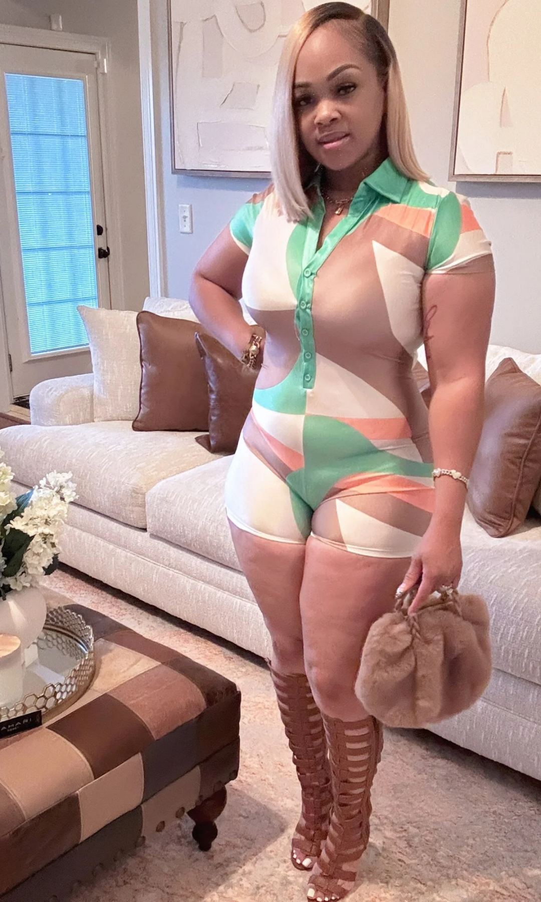 Front-facing view of a woman in a pastel green, peach, and beige patterned romper with a button-up collared neckline, accessorized with a tan faux-fur bag and brown gladiator heels.
