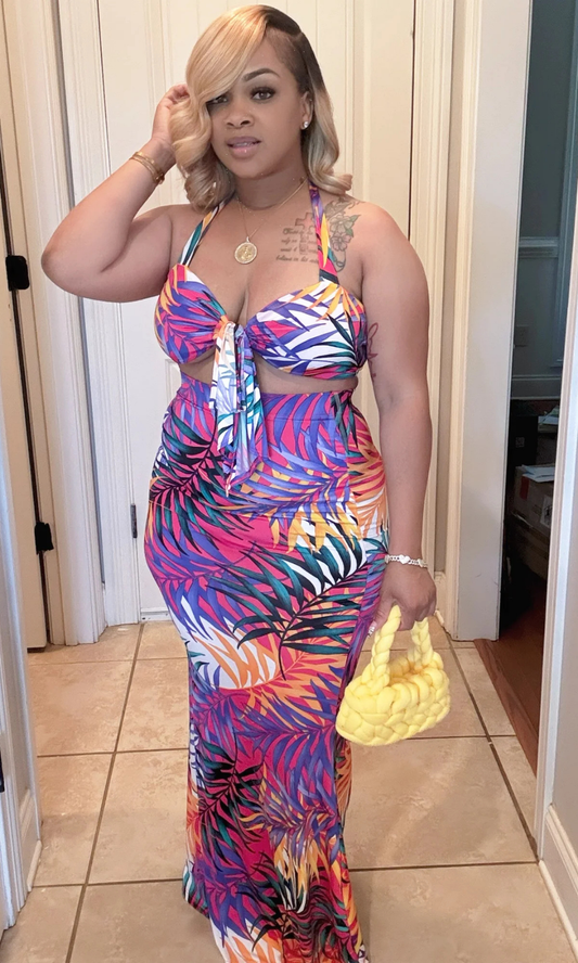 Front view of The Tropics Skirt Set featuring a halter crop top with a center tie and matching high-waisted, floor-length skirt in a bright tropical palm leaf print of pink, purple, yellow, orange, teal, and white. Model holds a small braided yellow handbag and wears gold jewelry.