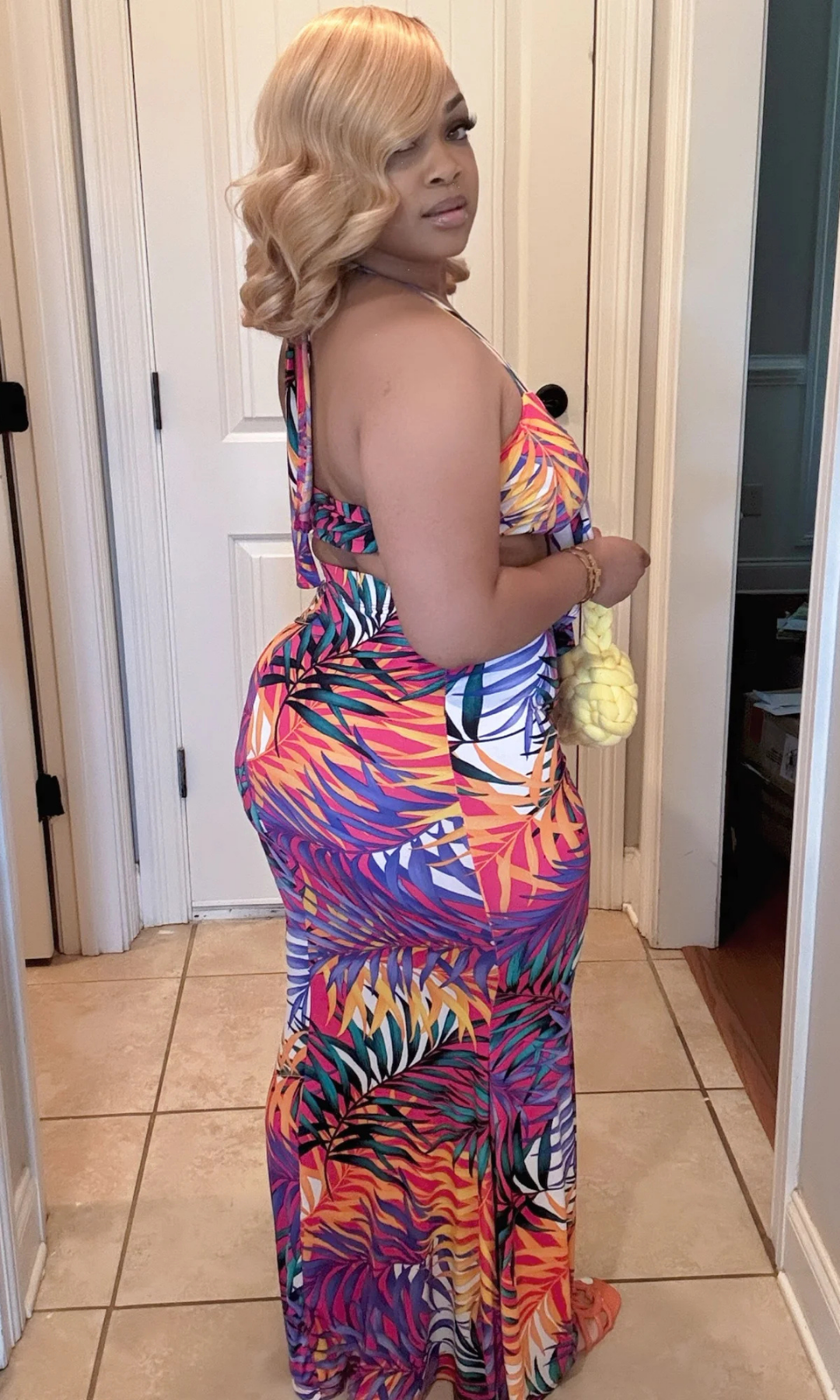 Side-back view of The Tropics Skirt Set showing the halter tie-back top and curve-hugging maxi skirt in the same bold tropical palm leaf print, paired with a braided yellow handbag.