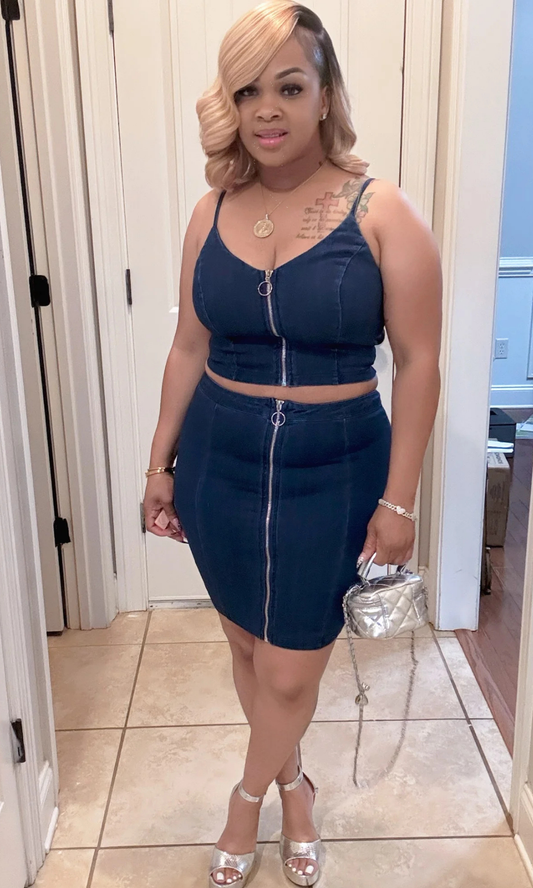 Front view of model wearing the Unzip Me Jean Set – a dark wash two-piece denim outfit with a sleeveless crop top and high-waisted skirt, both featuring bold silver front zippers, paired with silver heels and a quilted mini bag.