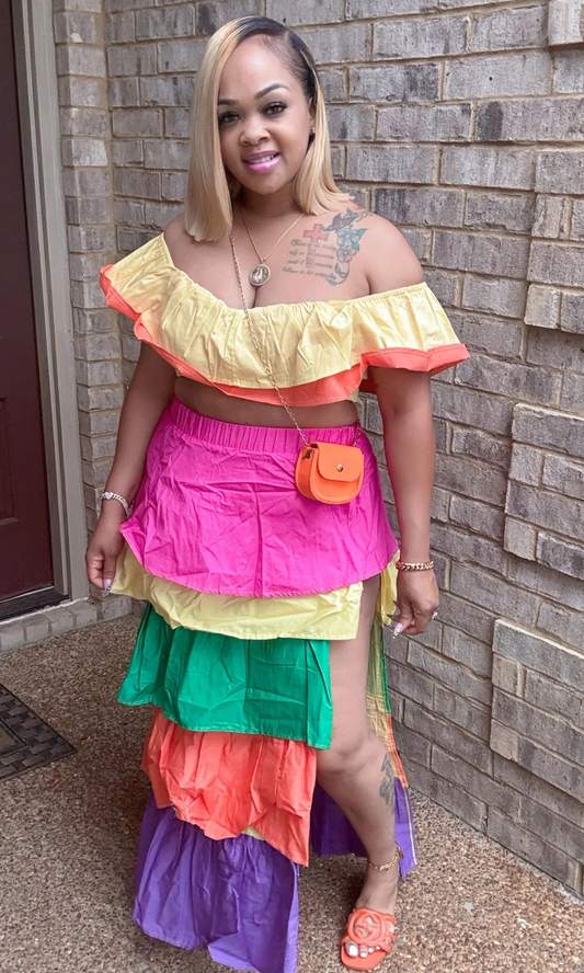 Front View – Woman wearing colorful 2-piece set with ruffled off-shoulder yellow and coral crop top, tiered maxi skirt in hot pink, yellow, green, orange, and purple, and high side slit.
