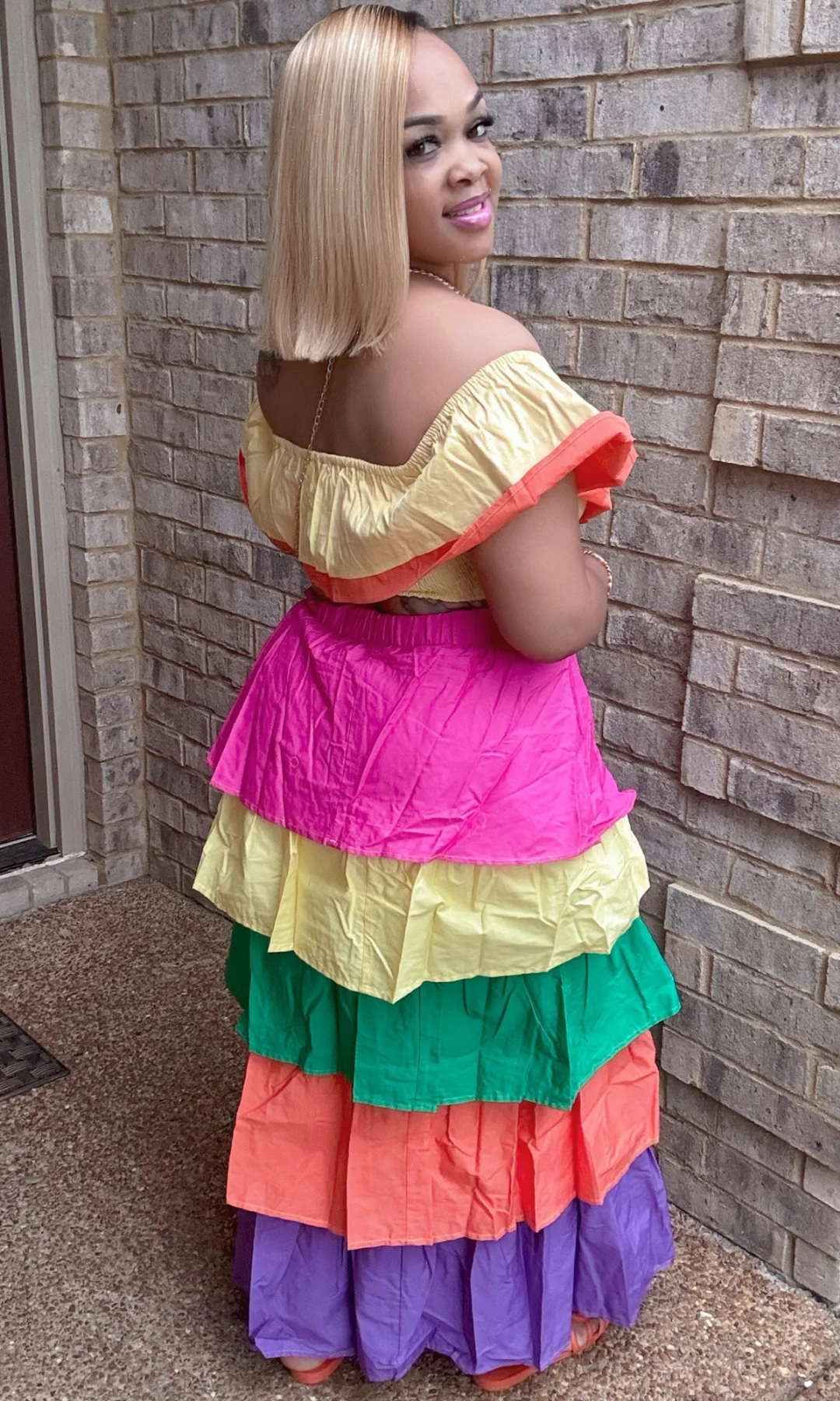 Back View – Woman showing back of colorful 2-piece set with layered ruffled off-shoulder crop top and tiered maxi skirt in vibrant tropical shades, standing against brick wall.

