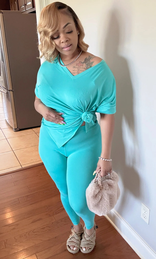 Woman wearing the V-Neck Ice Blue 2-Piece Set, featuring an oversized short-sleeve V-neck top knotted at the waist and matching fitted leggings, paired with rope-style slide sandals, a fuzzy beige handbag, and diamond jewelry, standing indoors on wooden flooring.