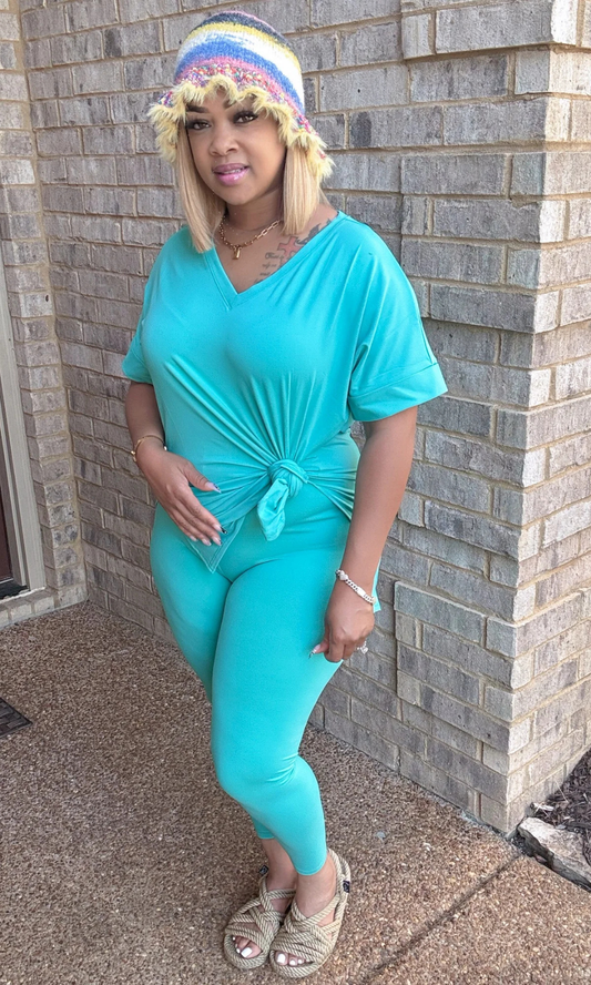 Woman wearing the V-Neck Turquoise 2-Piece Set, styled with a knotted oversized V-neck top and matching fitted leggings in a vivid turquoise shade, accessorized with a multicolored knit bucket hat, rope sandals, gold bracelet, silver chain bracelet, and sleek blonde bob, standing against a brick wall.

