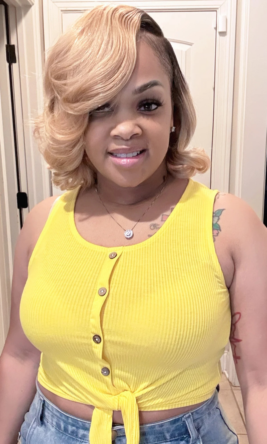 Woman wearing a sleeveless yellow ribbed crop top with front buttons and a tie-front hem, paired with high-waisted denim shorts, smiling indoors.

