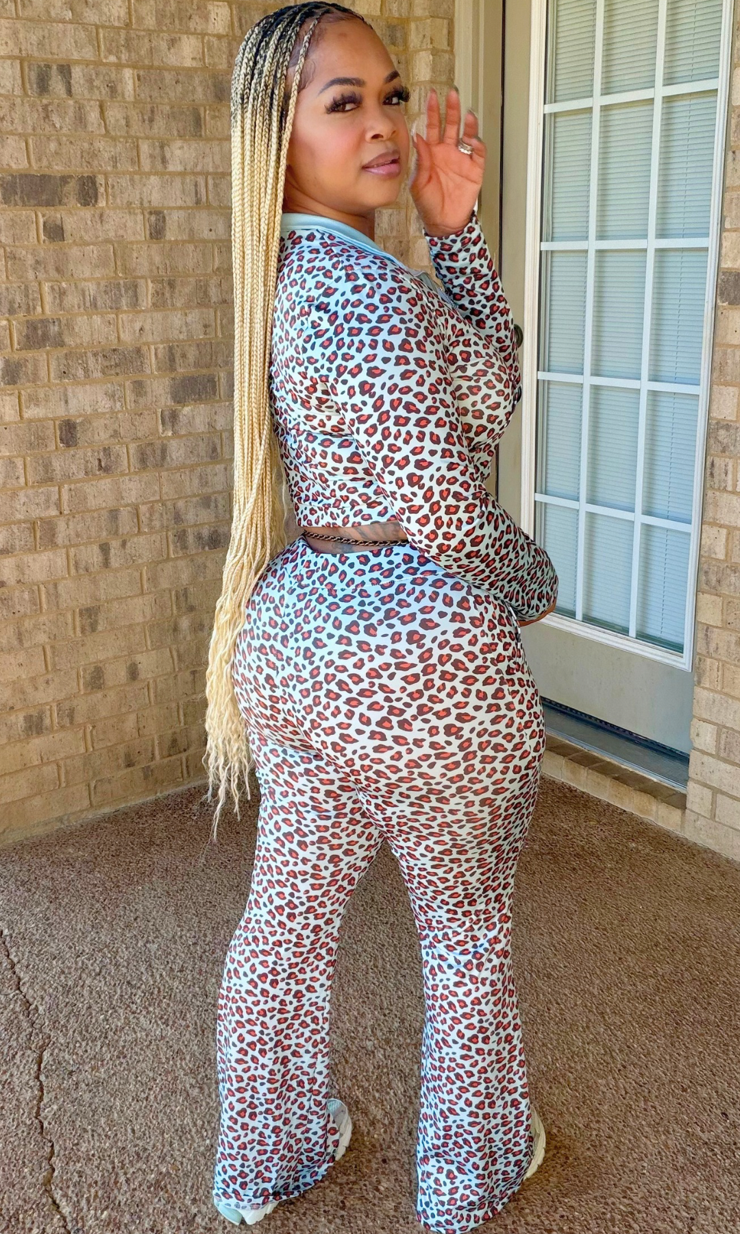 A woman wearing a matching two-piece leopard print jumpsuit set, standing on a concrete porch next to a brick wall and glass door.