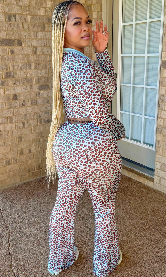 A woman wearing a matching two-piece leopard print jumpsuit set, standing on a concrete porch next to a brick wall and glass door.