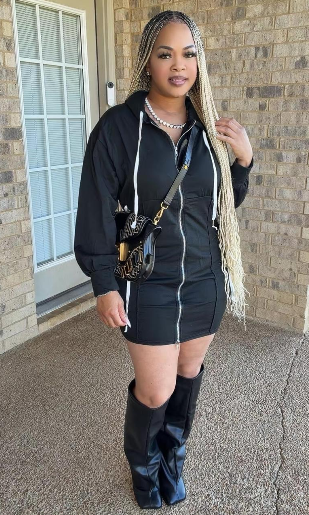 Model wearing the Bow Tie Hoodie Dress Black, featuring a full-length front zipper, hood with white drawstrings, long sleeves, and a fitted mini-length silhouette, styled with black knee-high boots.