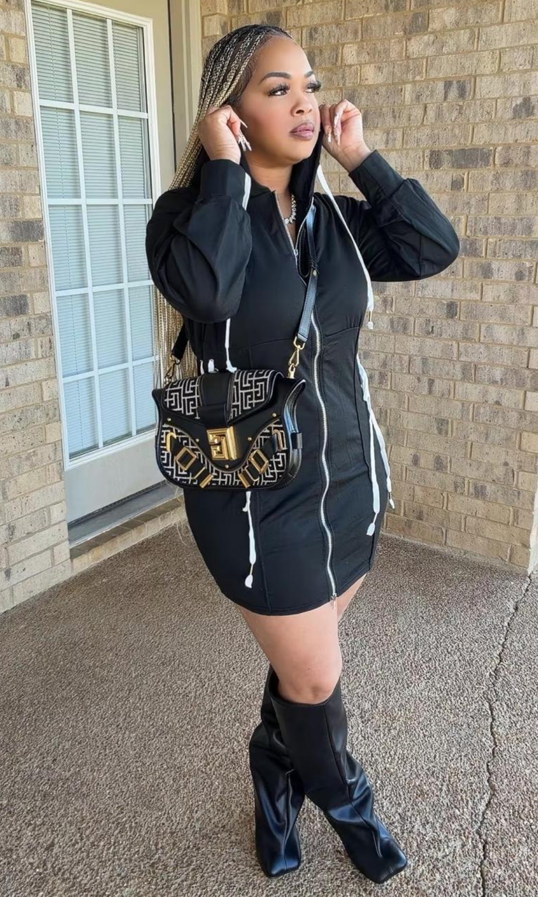 A woman wearing the Bow Tie Hoodie Dress Black, featuring a full-length front zipper, hood with white drawstrings, long sleeves, and a fitted mini-length silhouette, styled with black knee-high boots.