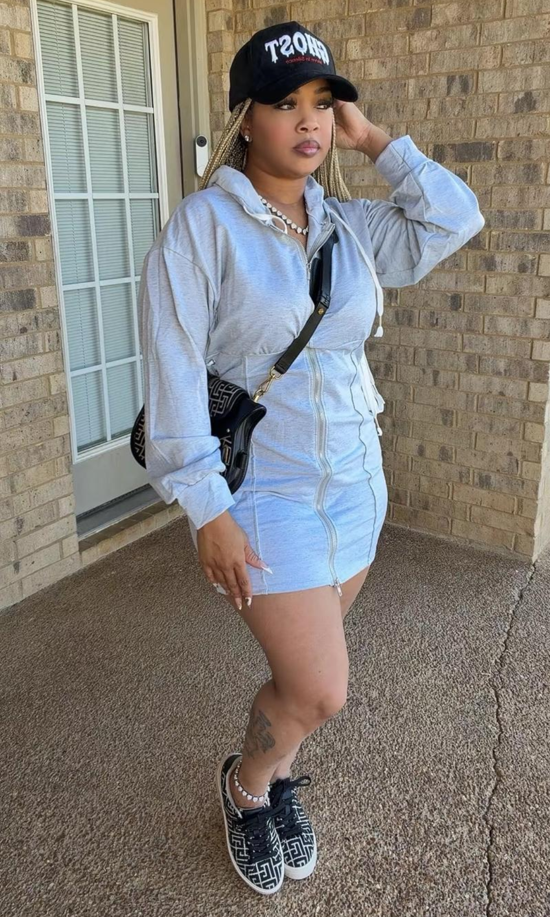 Model wearing the Bow Tie Hoodie Dress Gray, featuring a heather gray mini-length silhouette, front zipper, attached hood with white drawstrings, long sleeves, and lace-up side details, styled with sneakers and a black baseball cap.