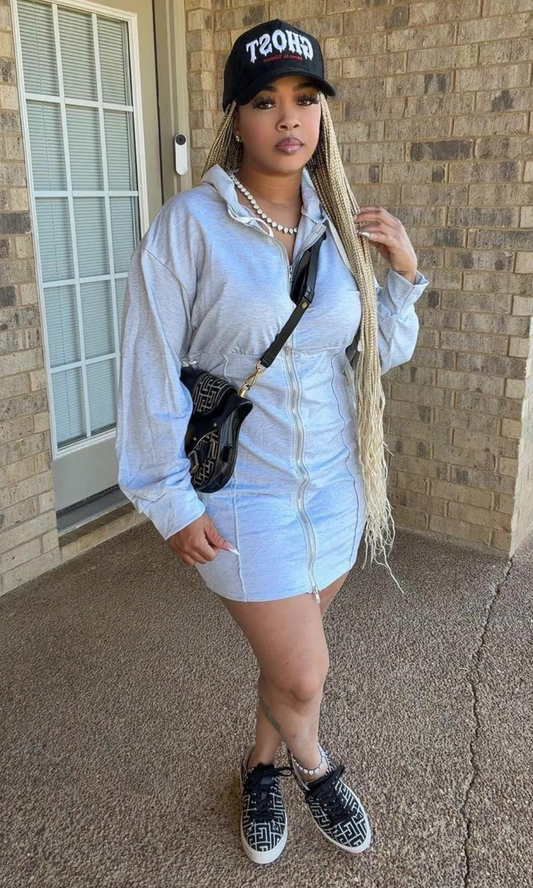 The model is wearing the Bow Tie Hoodie Dress Gray, featuring a heather gray mini-length silhouette, front zipper, attached hood with white drawstrings, long sleeves, and lace-up side details, styled with sneakers and a black baseball cap.