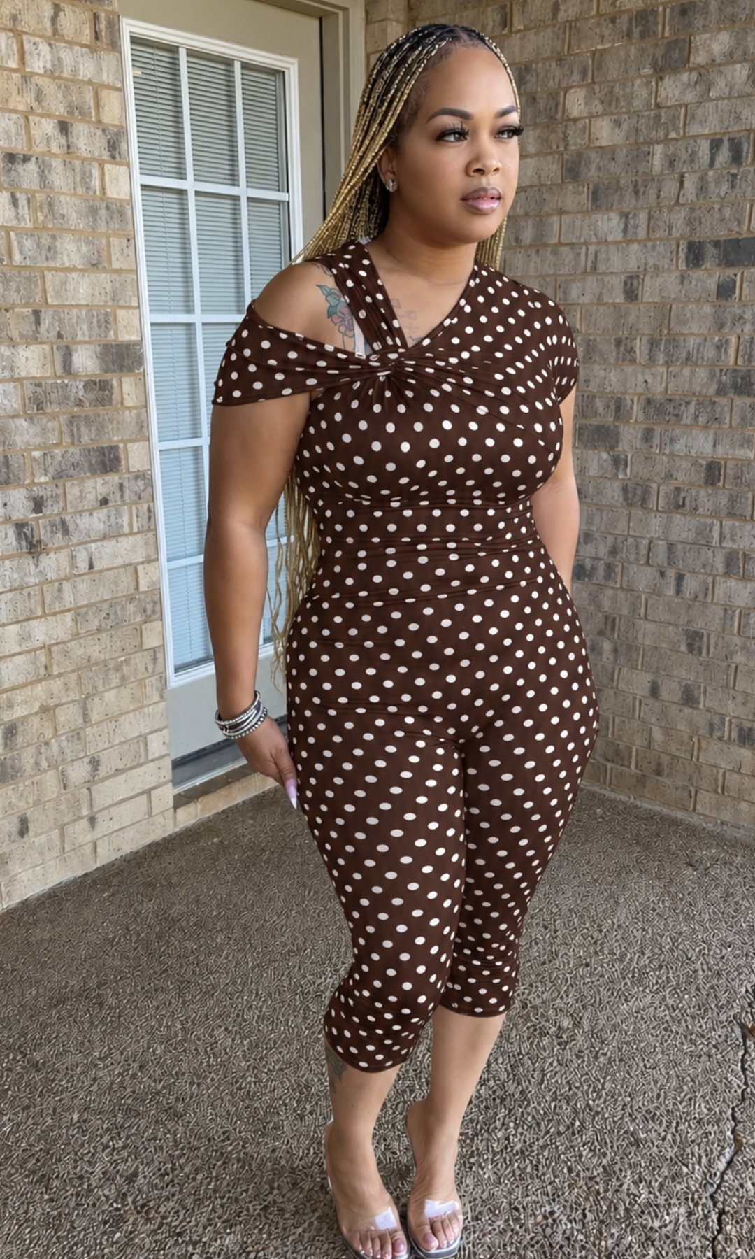 Woman modeling the Brown Polka Babe Jumpsuit featuring a chocolate brown and white polka dot print, twisted asymmetrical neckline, off-shoulder detail, and fitted capri-length legs.